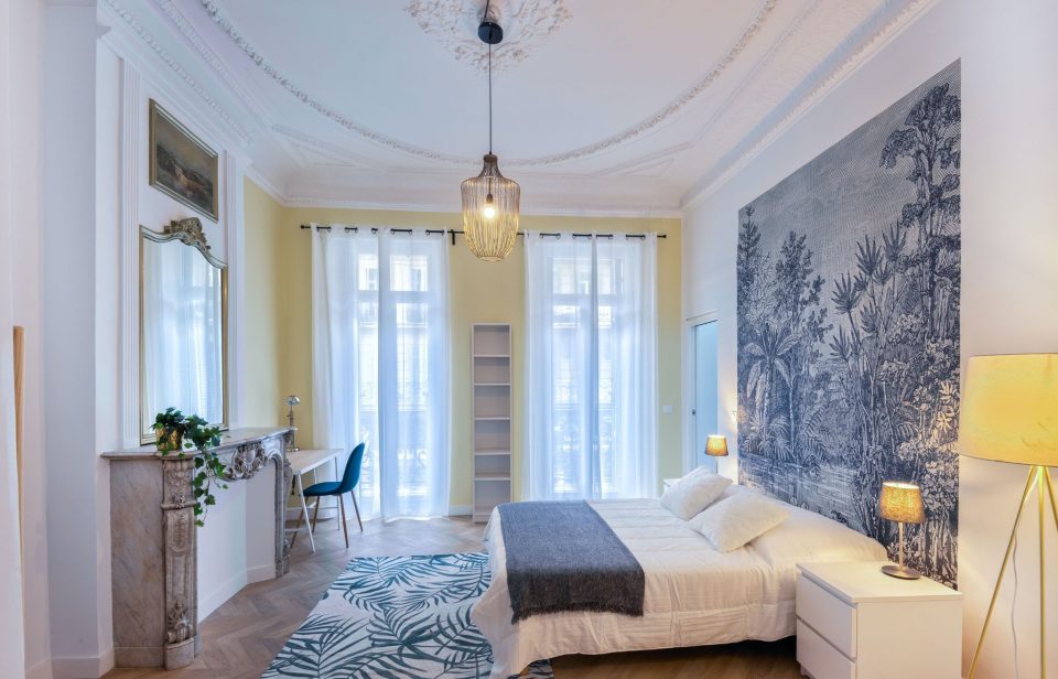 Chambre avec mur jaune pâle et papier peint graphique dans un appartement ancien à haute hauteur sous plafond