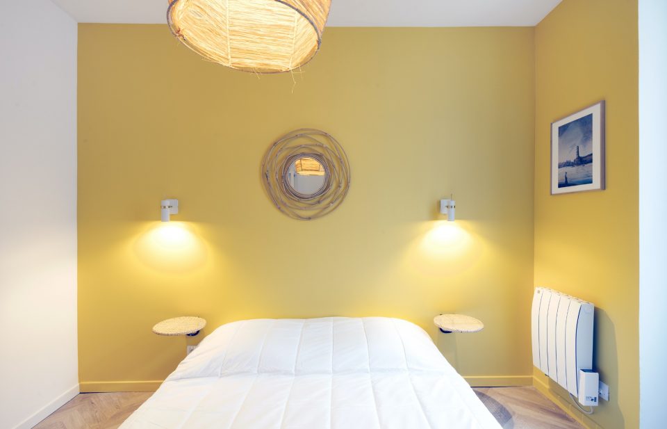 Chambre avec mur jaune moutarde et appliques symétriques dans un appartement locatif rénové