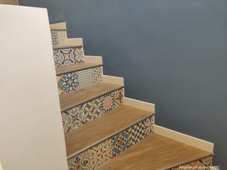 Escalier intérieur avec marches en bois et contremarches décorées de carreaux colorés à motifs, contre un mur bleu uni.
